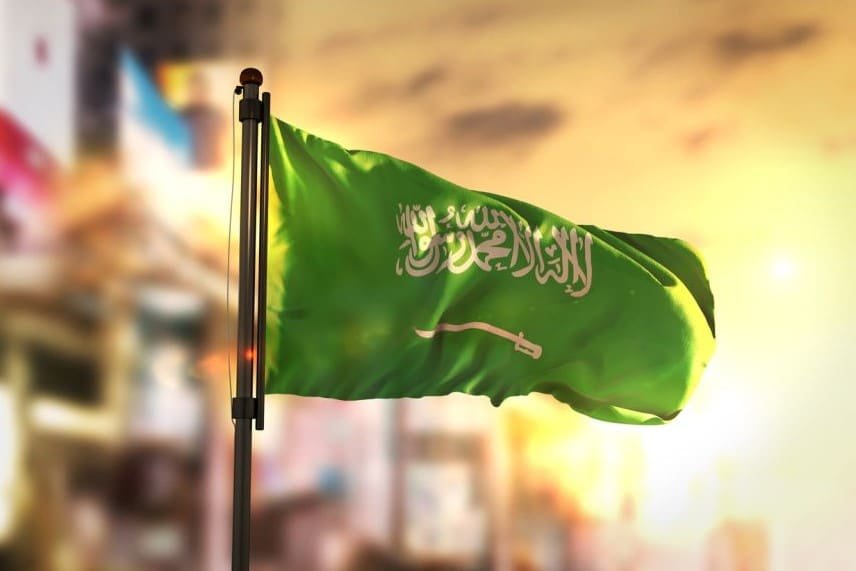 The flag of Saudi Arabia waving proudly on a flagpole, backlit by warm, golden sunlight.